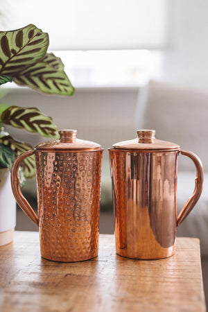 Kopero Copper Jug, a smooth and a hammered jug side by side on a wooden table, with a plant and a sofa behind