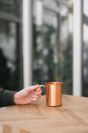 Kopero Copper Mug Smooth, on a wooden table, with a hand on the handle