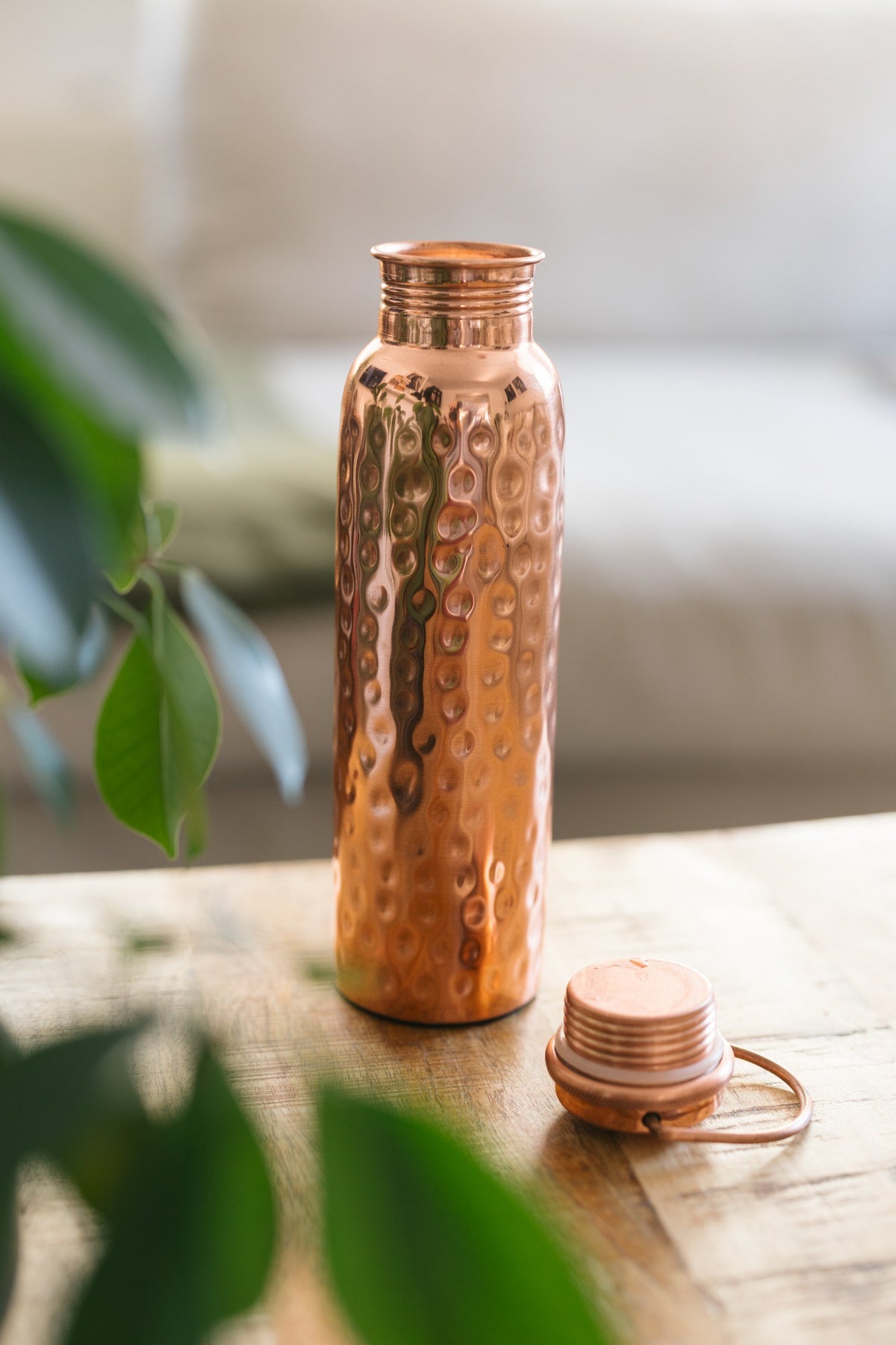Kopero Copper Water Bottle Hammered, standing on a wooden table in front of a sofa, with the loose cap next to it