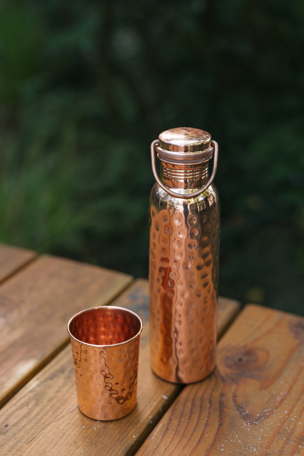 Kopero Copper Water Bottle Hammered, on a wooden garden table next to a hammered cup