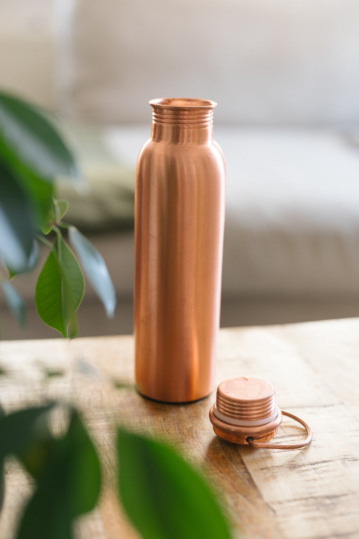 Kopero Copper Water Bottle Smooth, standing on a wooden table in front of a sofa, with the loose cap next to it