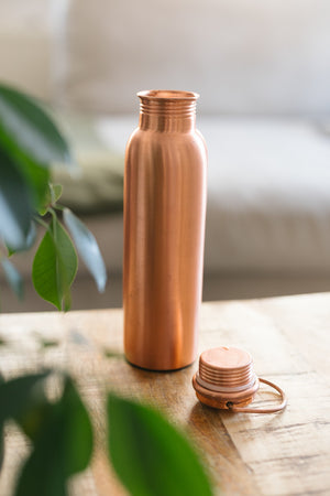 Kopero Copper Water Bottle Smooth, standing on a wooden table in front of a sofa, with the loose cap next to it